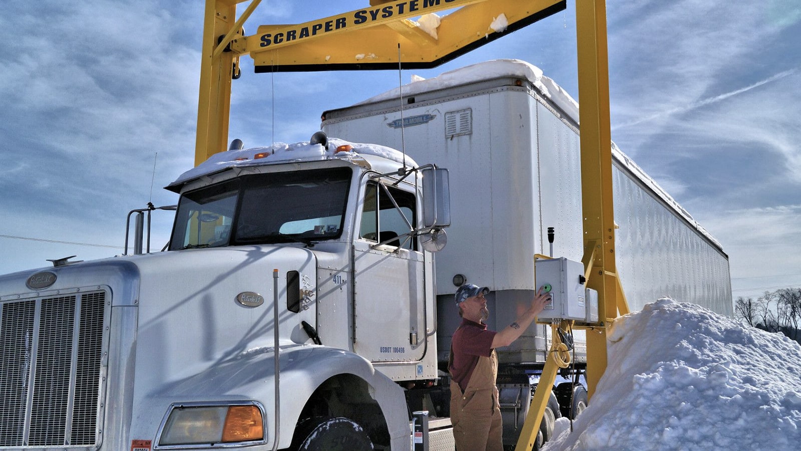 A truck and trailer driving under an automated snow removal scraper system.